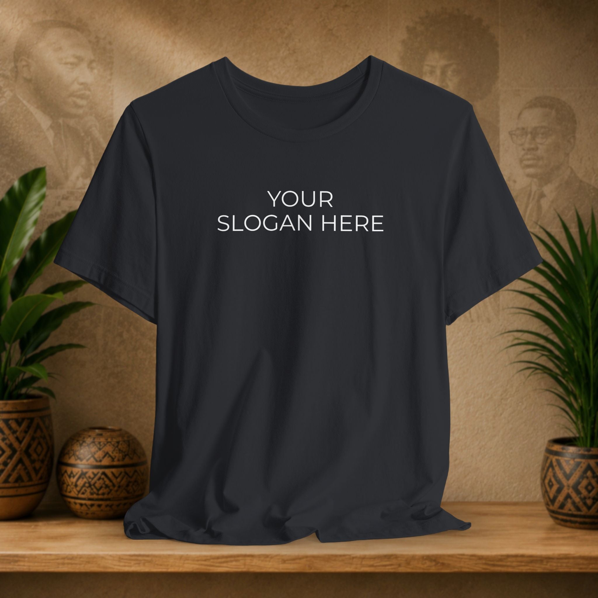 A black t-shirt with 'YOUR SLOGAN HERE' printed in white text on the chest, displayed on a wooden shelf with decorative plants and textured background.