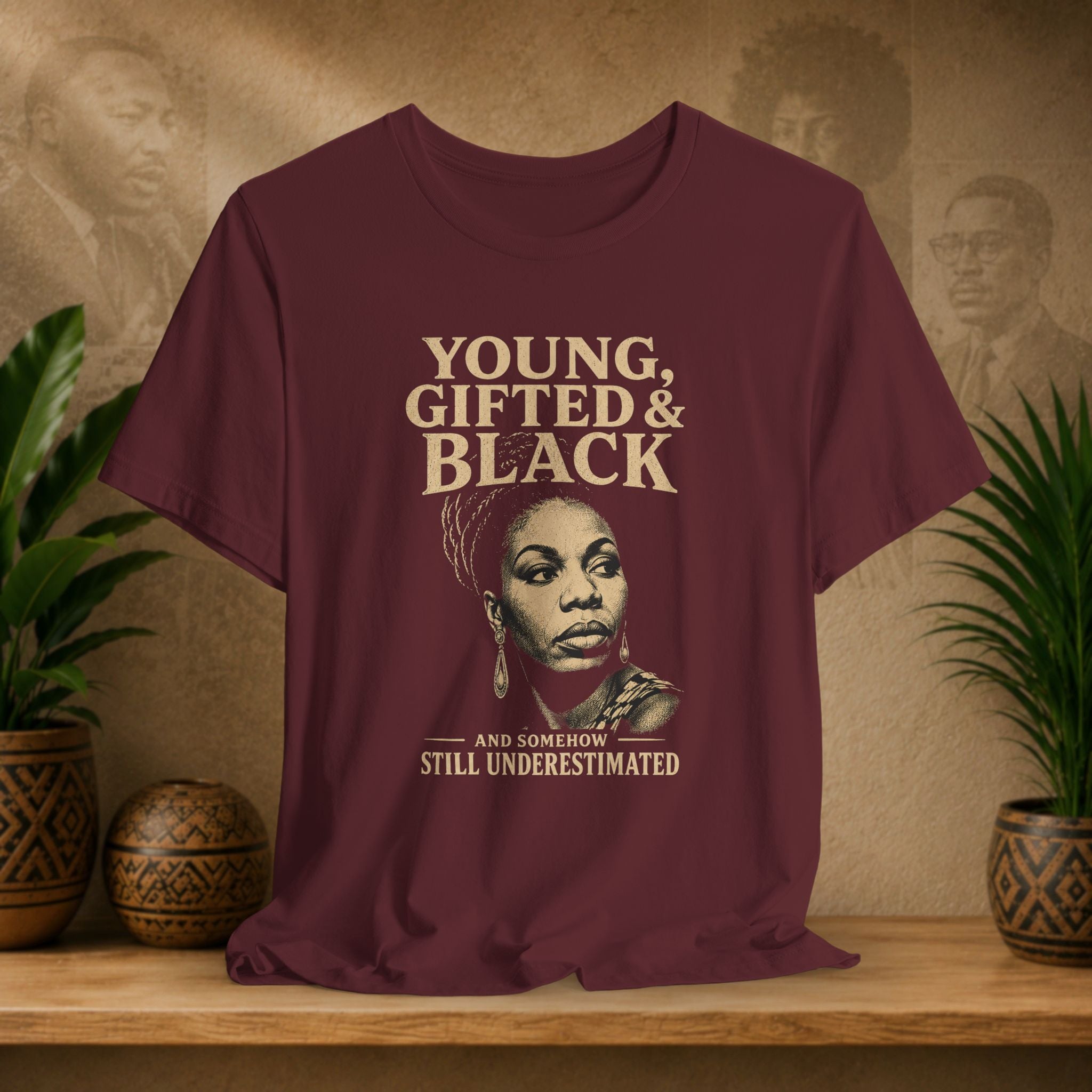 A maroon short-sleeve graphic T-shirt displayed on a wooden shelf, flanked by potted green plants against a textured beige wall with faint portraits. The shirt features a large front graphic with the text 'YOUNG, GIFTED & BLACK' and 'AND SOMEBOW STILL UNDERESTIMATED' around an illustrated portrait.