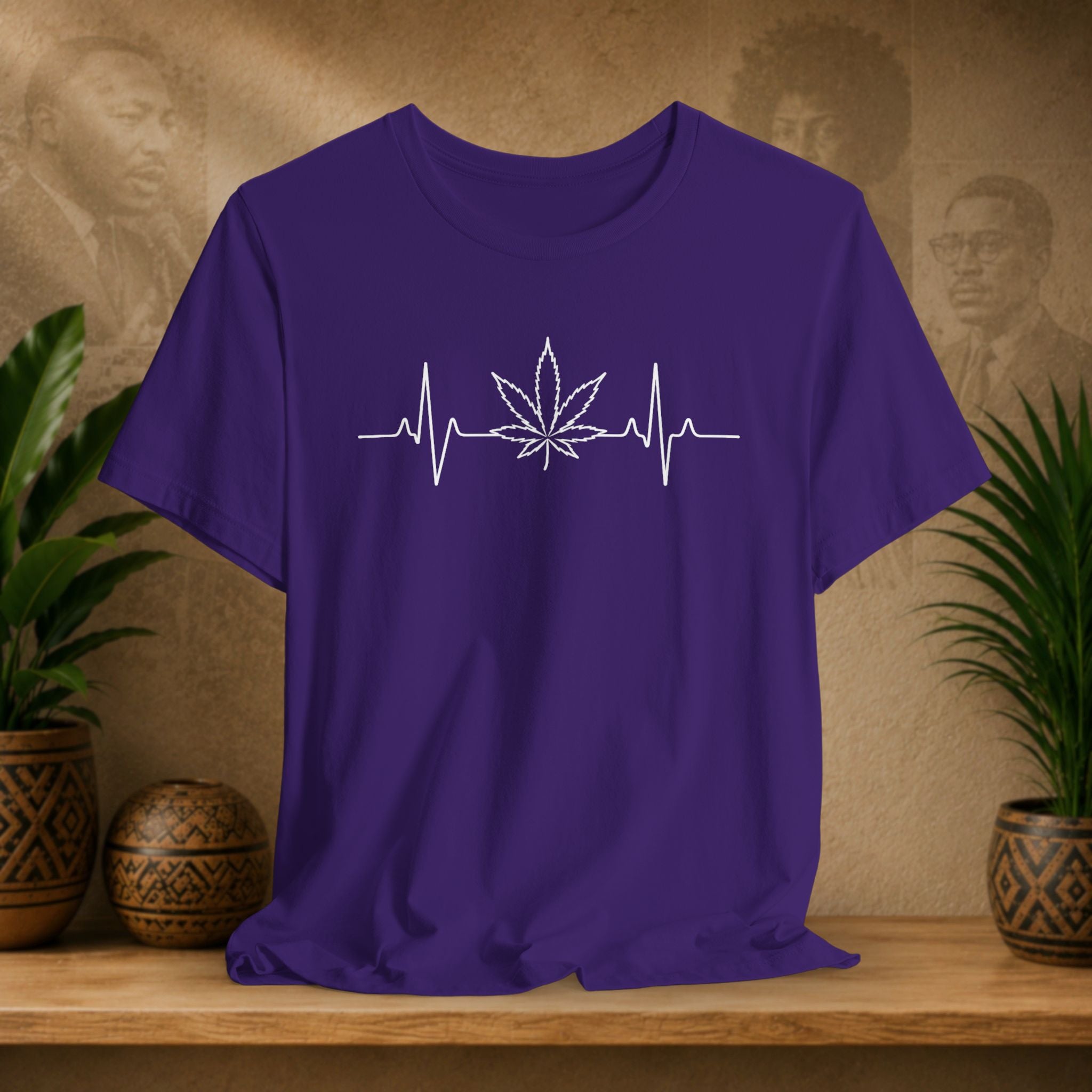 Purple t-shirt with a white cannabis leaf and heartbeat line design on a wooden surface with plants and a textured wall background.