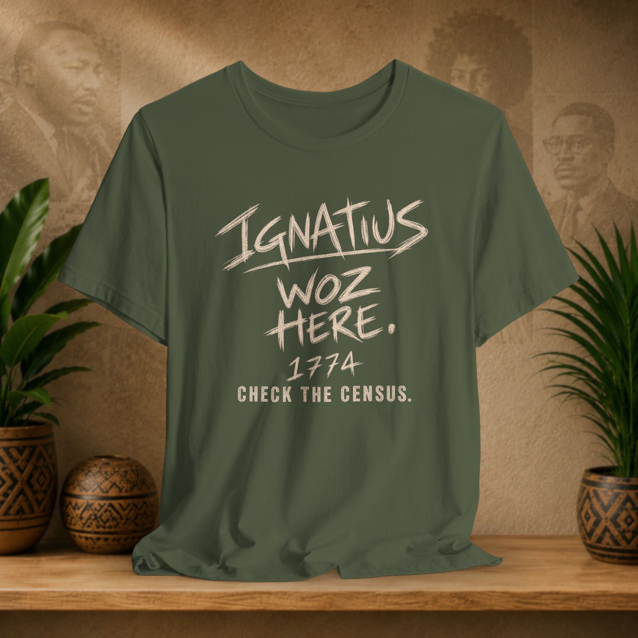 An olive green graphic t-shirt displayed on a wooden shelf, flanked by potted plants. The shirt features a distressed white graphic print that reads 'Ignatius Woz Here.