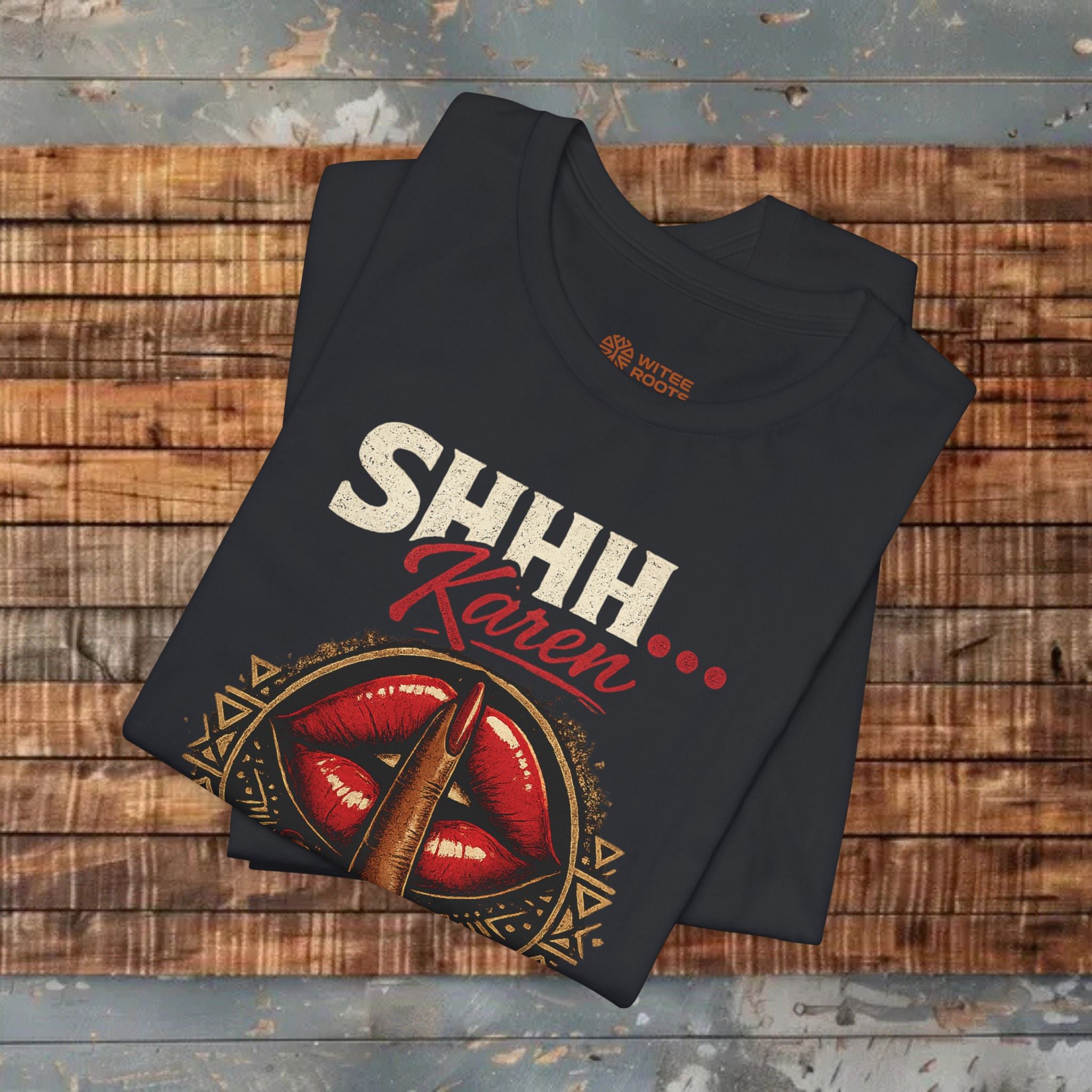 A folded black graphic T-shirt displayed on a rustic wooden plank surface. The shirt features a graphic print with the text 'SHHH Karen' in white and red, above an illustration of red lips with a bullet embedded in them and gold geometric details.