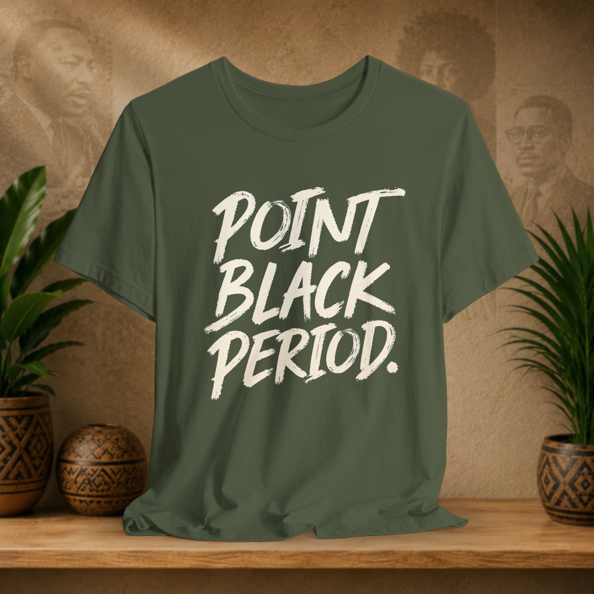 An olive green short-sleeve t-shirt with the text 'POINT BLACK PERIOD.' printed in a white brush-stroke font, displayed on a wooden shelf with potted plants against a textured beige background.