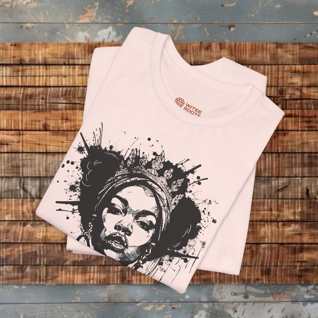 A folded light pink t-shirt with a black and white graphic print of a woman's face surrounded by paint splatters, displayed on a rustic wooden plank surface.