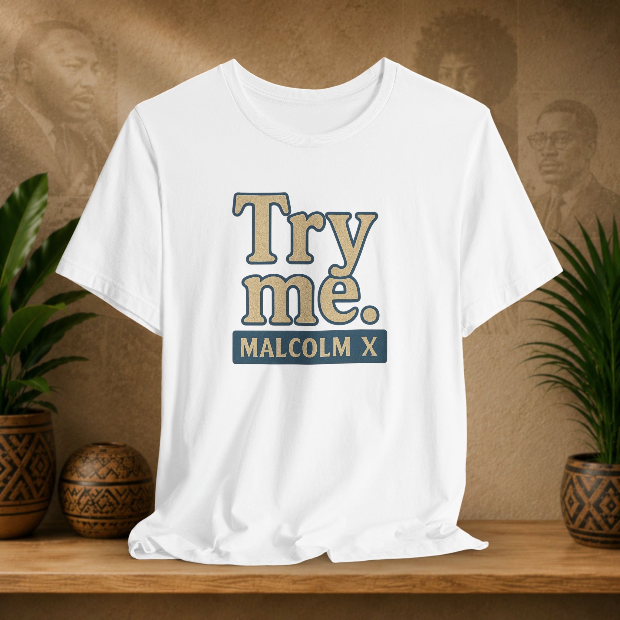 White t-shirt with 'Try me. MALCOLM X' text on a wooden shelf with plants and a textured wall background.