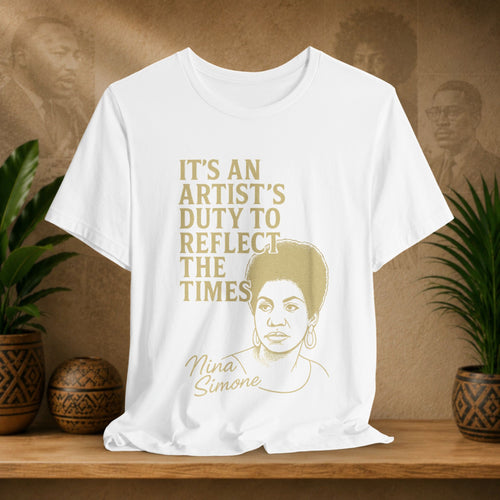 White t-shirt with gold text and Nina Simone's portrait on a wooden surface with plants and a brown background.