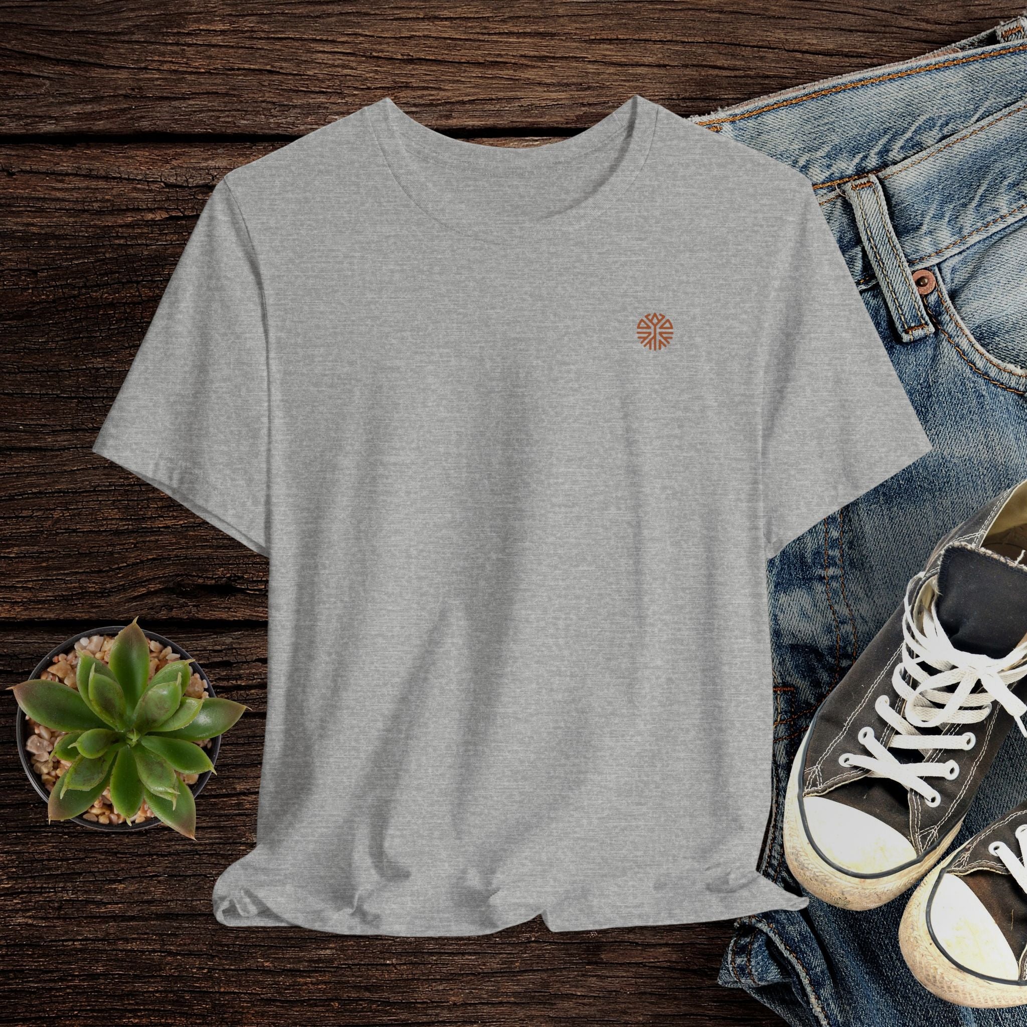 Gray t-shirt with a small logo on a wooden surface with jeans and sneakers.