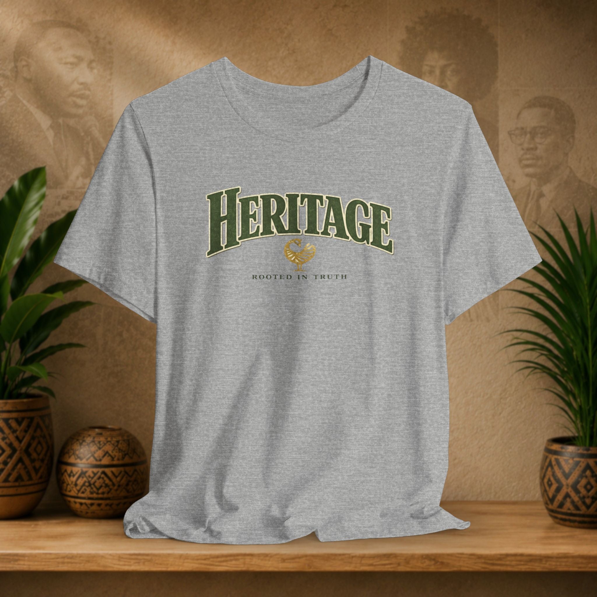 Gray t-shirt with 'HERITAGE' printed on it, displayed on a wooden shelf with plants and a decorative item.