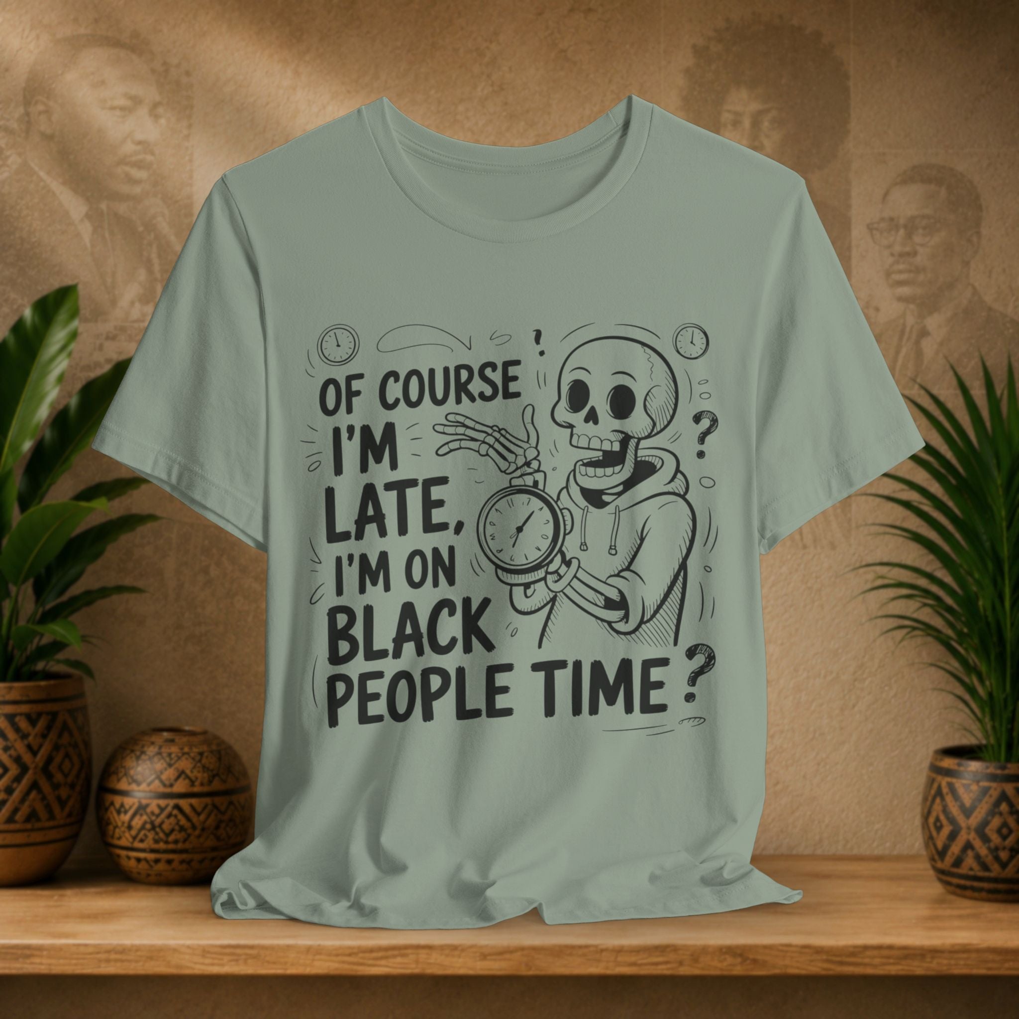 A sage green graphic t-shirt displayed on a wooden shelf, flanked by potted plants. The shirt features a black line art graphic of a skeleton in a hoodie holding a clock, with the text 'OF COURSE I'M LATE, I'M ON BLACK PEOPLE TIME ?'.