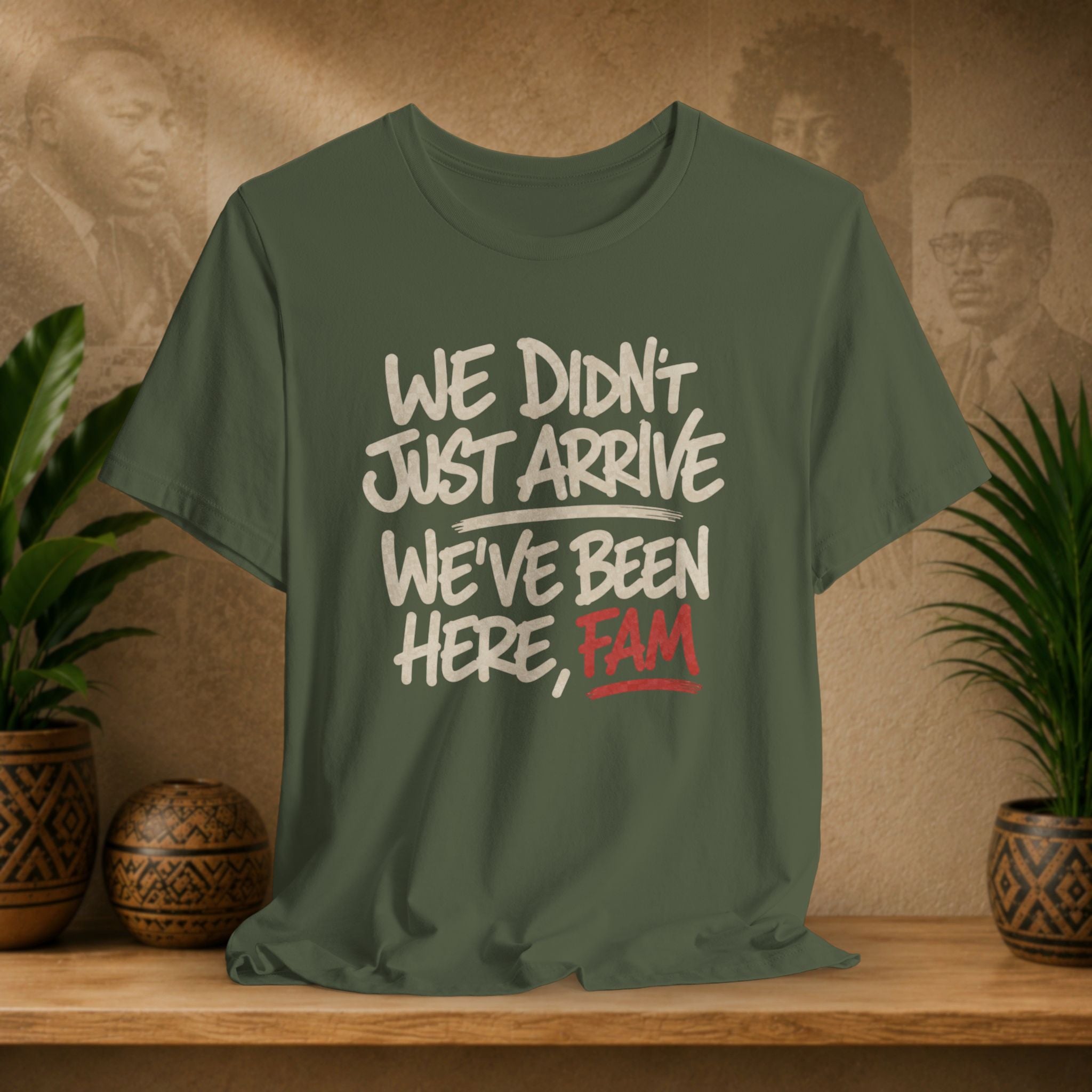 An olive green graphic t-shirt displayed on a wooden shelf, flanked by potted plants. The shirt features a distressed white and red text print that reads 'WE DIDN'T JUST ARRIVE WE'VE BEEN HERE, FAM'.