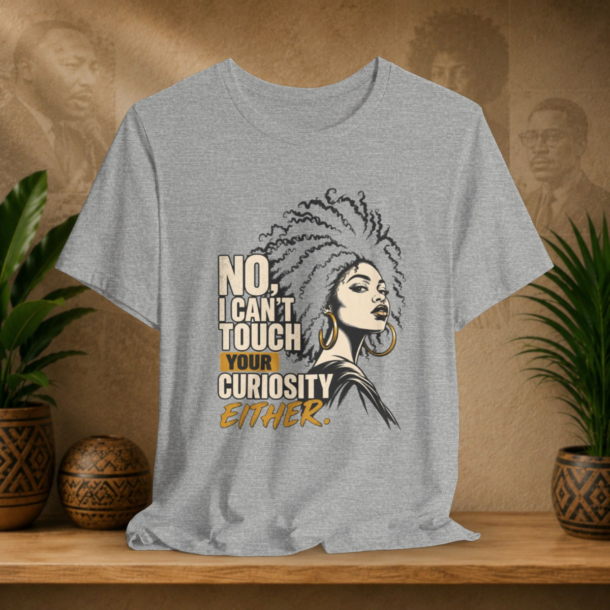 A heather gray graphic t-shirt displayed on a wooden shelf. The shirt features a stylized illustration of a Black woman with an afro and gold hoop earrings next to the text 'NO, I CAN'T TOUCH YOUR CURIOSITY EITHER.