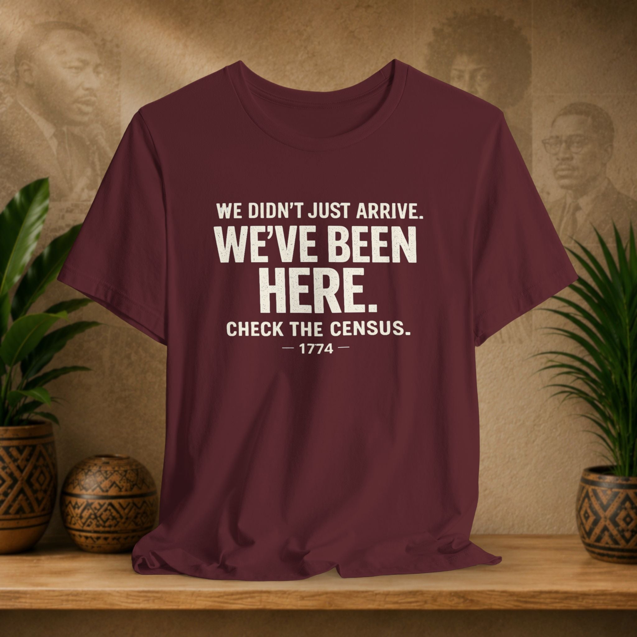 Maroon t-shirt with white text on a wooden surface with plants and a textured wall background
