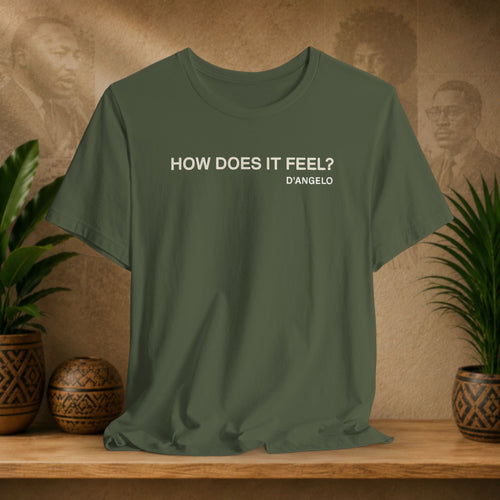 Green t-shirt with 'HOW DOES IT FEEL? D'ANGELO' text on a wooden surface with decorative plants and background.