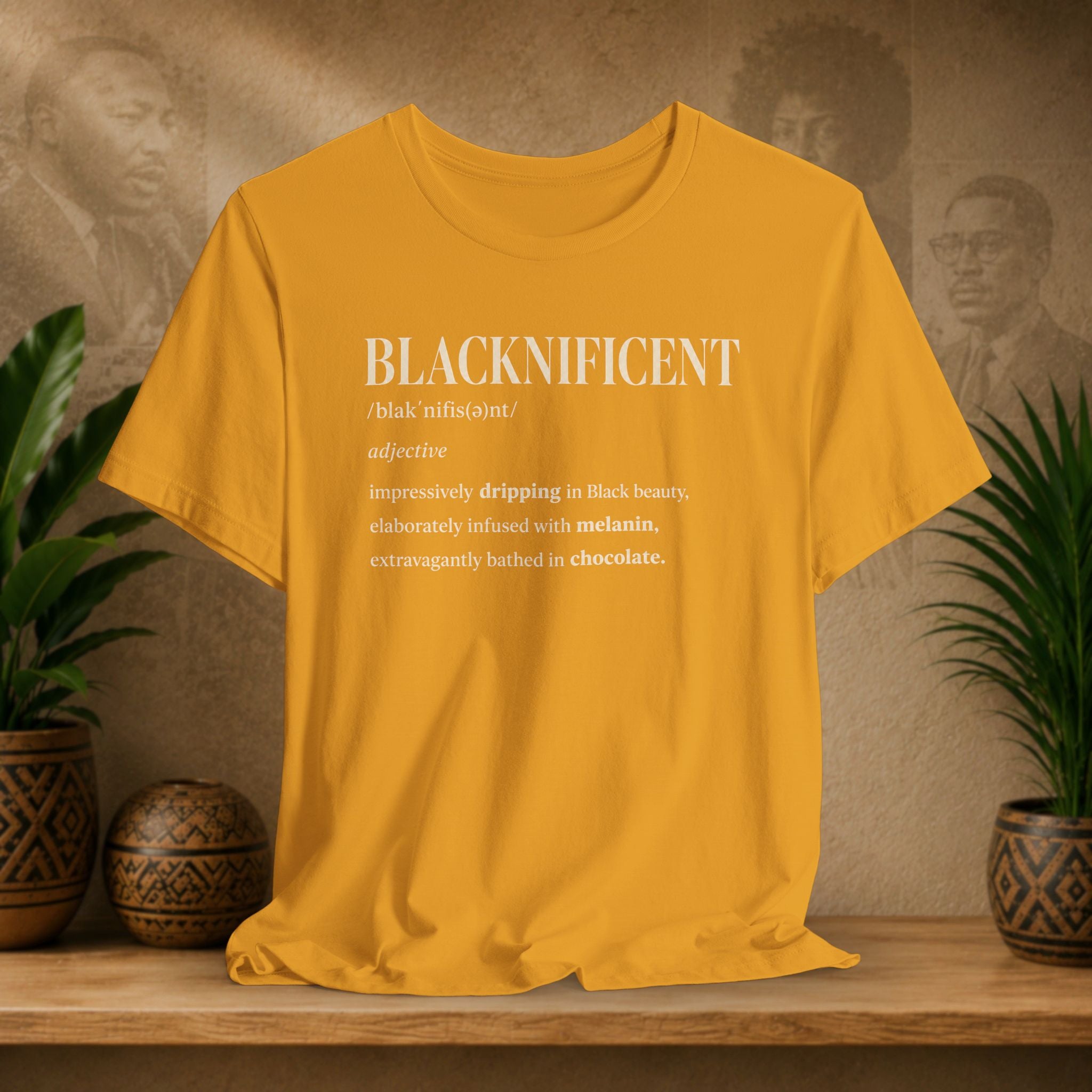 A mustard yellow graphic t-shirt displayed on a wooden shelf, flanked by potted green plants and decorative ceramic pots against a textured beige wall with vintage portraits.