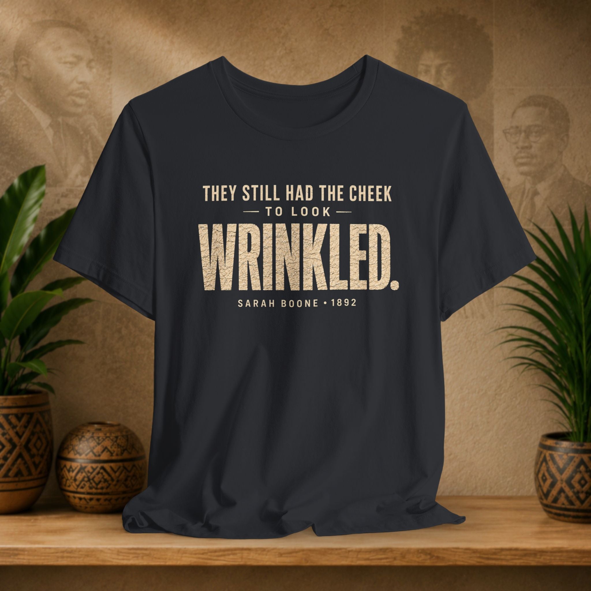 Black t-shirt with white text on a wooden surface with plants and a textured wall background