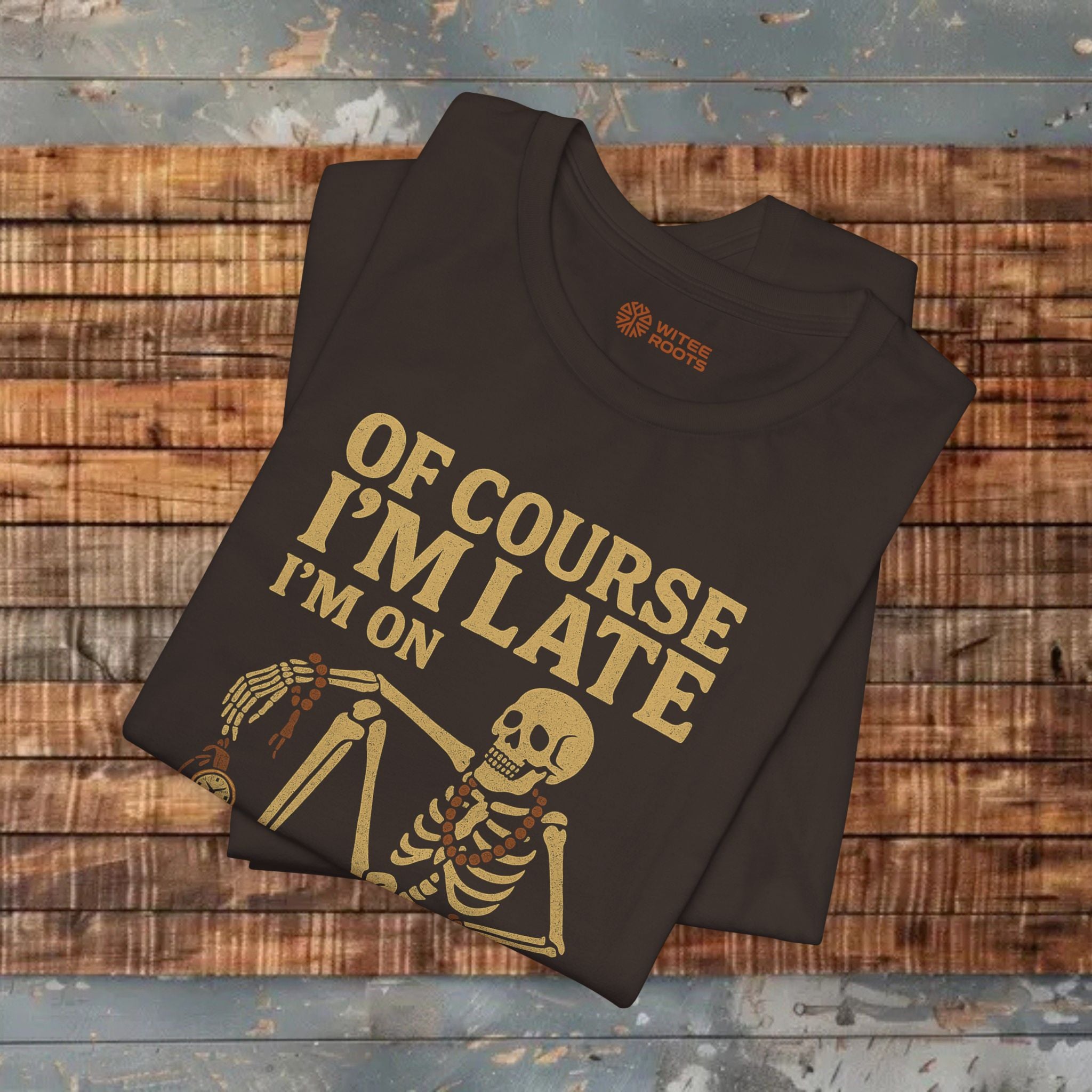 A folded brown graphic T-shirt displayed on a rustic wooden plank surface. The shirt features a gold-colored skeleton illustration and the text 'OF COURSE I'M LATE I'M ON'.