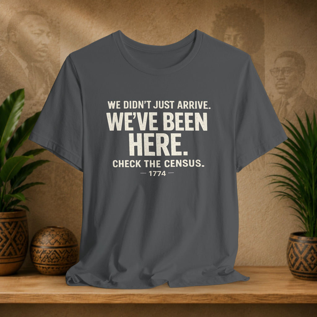 Grey t-shirt with white text on a wooden surface with plants and a textured wall background
