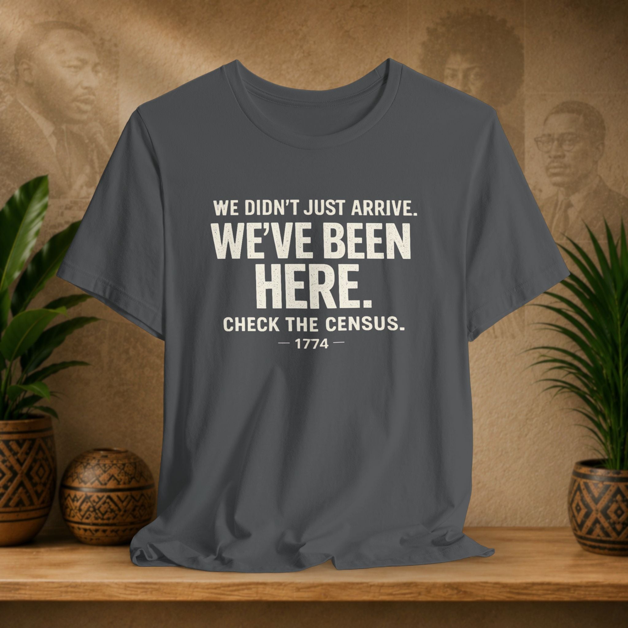 Grey t-shirt with white text on a wooden surface with plants and a textured wall background