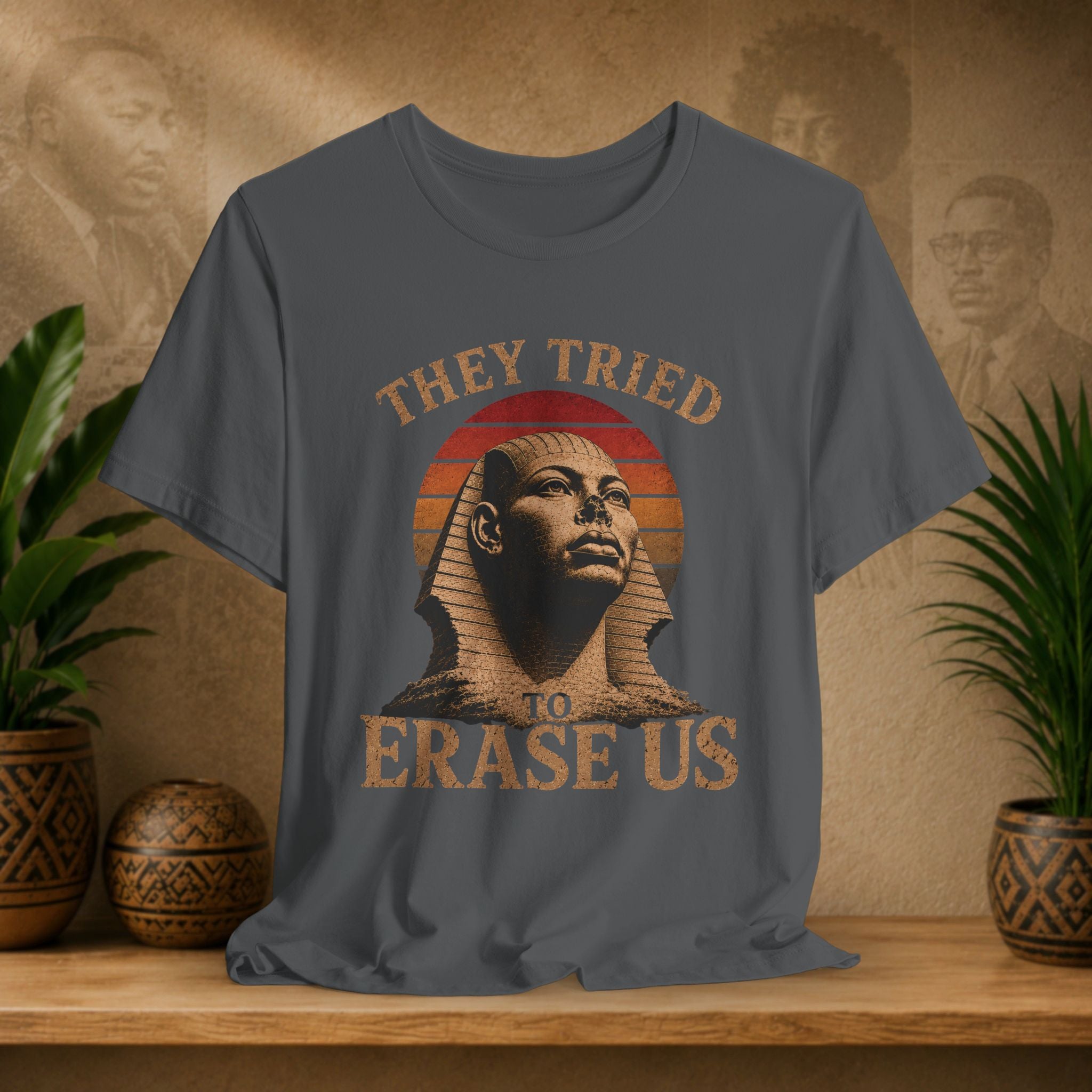 T-shirt with a design of a sphinx and text 'They Tried To Erase Us' on a wooden surface with decorative plants and background.