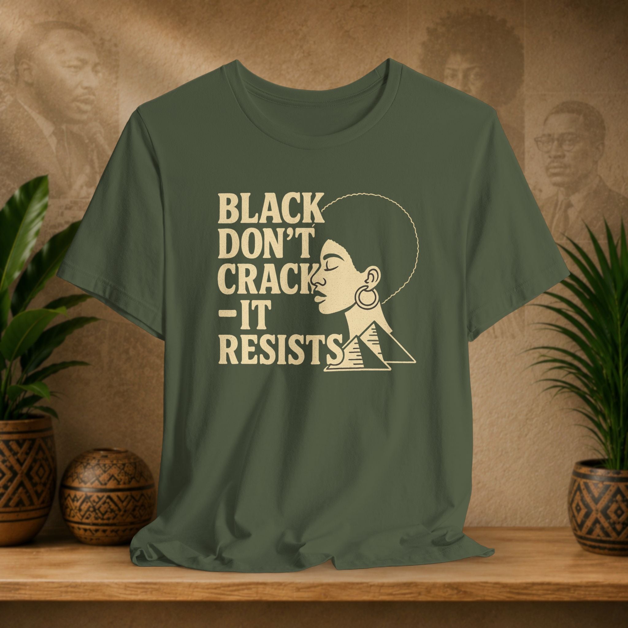 Green t-shirt with 'Black Don't Crack, It Resists' text and graphic on a wooden surface with a decorative background.