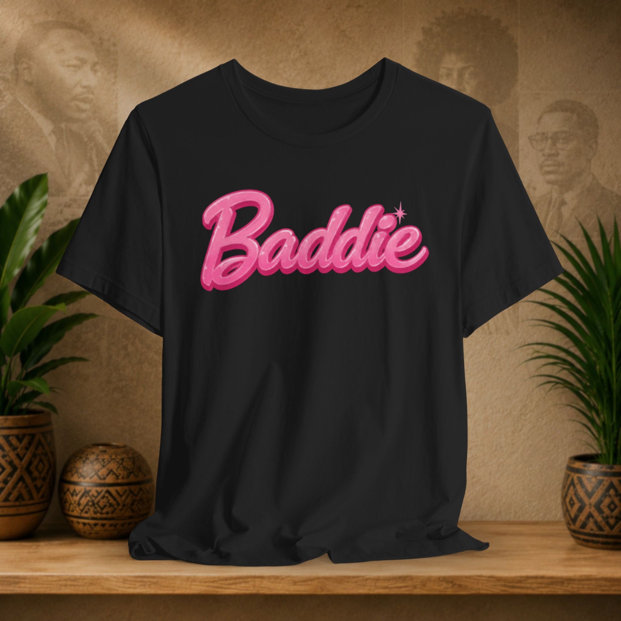 A black short-sleeve t-shirt with 'Baddie' printed in a pink 3D bubble font across the chest, styled on a wooden shelf with potted plants against a textured beige background.