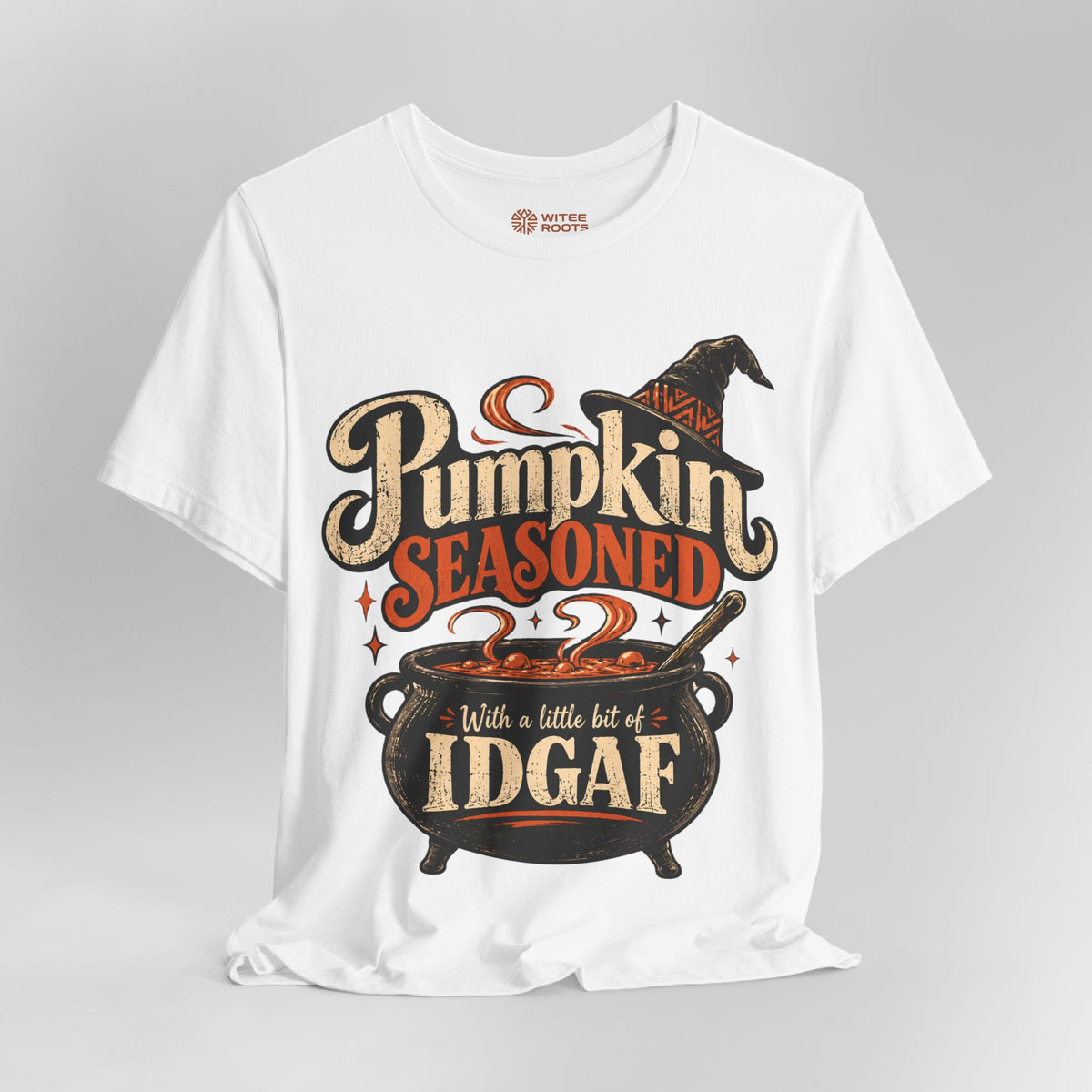 A white short-sleeve graphic t-shirt displayed on a neutral gray background. The shirt features a Halloween-themed graphic with the text 'Pumpkin Seasoned' and 'With a little bit of IDGAF' surrounding a cauldron and witch hat.