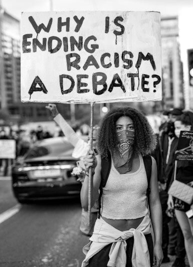 Person holding a protest sign asking 'Why is ending racism a debate?' in an urban setting.